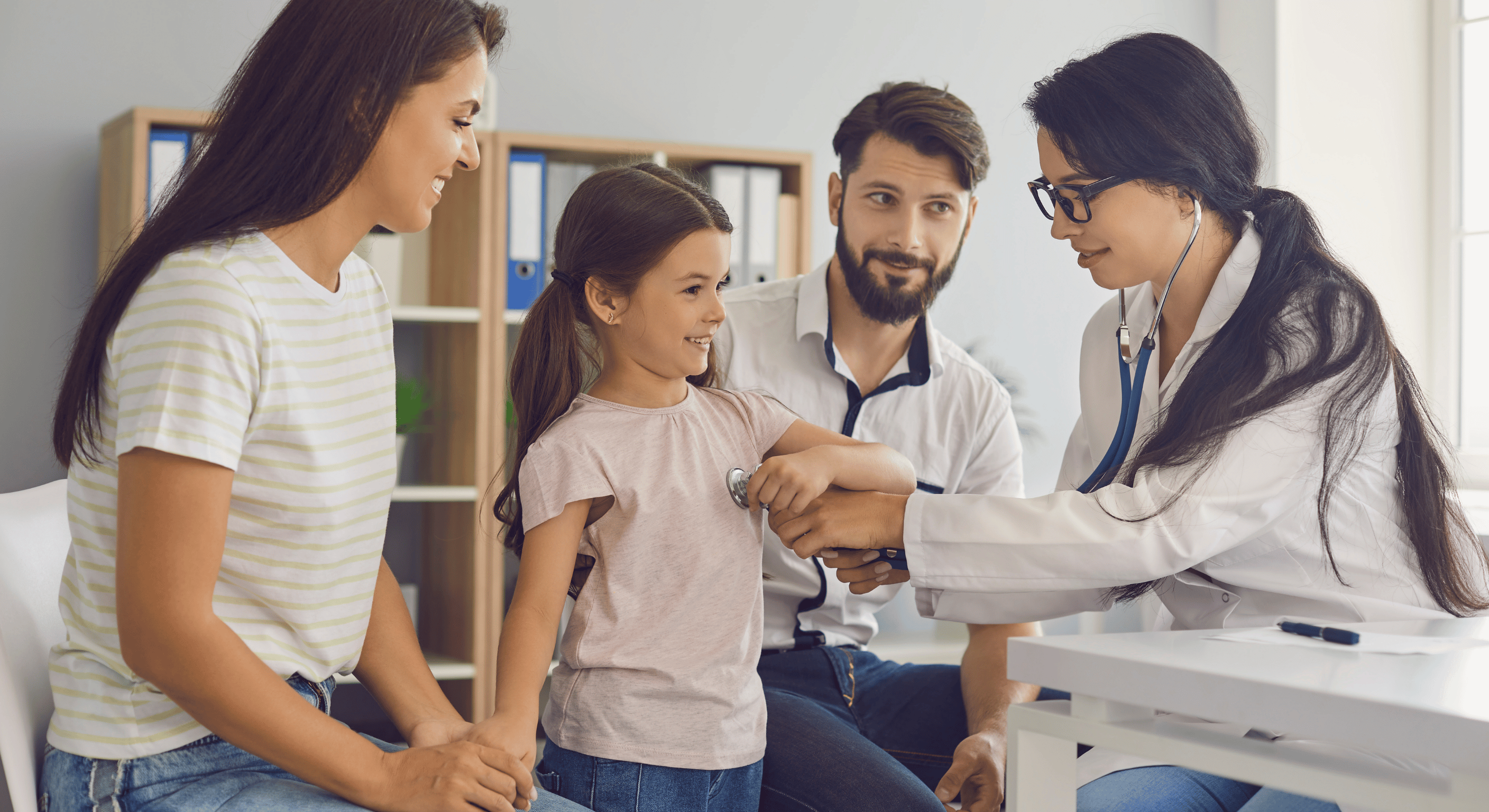 Doctor examining a smiling young girl with a stethoscope while her parents look on in a medical office